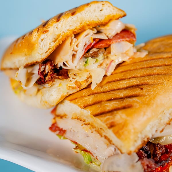 Two halves of a grilled panini sandwich filled with meat, cheese, and vegetables on a white plate.