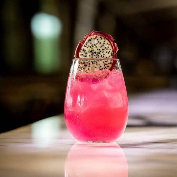 Pink cocktail with ice and a dragon fruit slice garnish on a reflective surface.