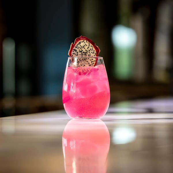 Vibrant pink cocktail with ice and a dragon fruit slice, reflecting on a polished surface.