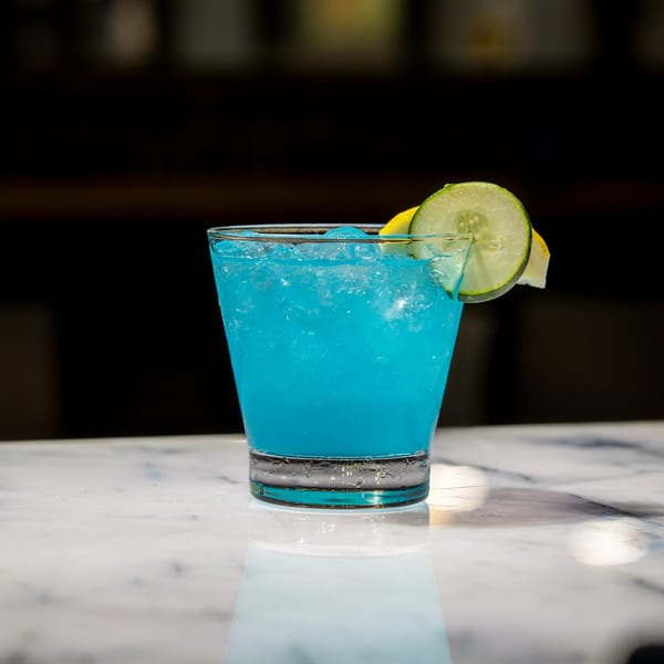 Vibrant blue cocktail with ice, garnished with a cucumber slice and lemon, on a marble surface.