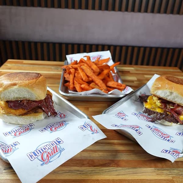 Two Clutch City Kitchen burgers with bacon and cheese, served with sweet potato fries on a wooden table.