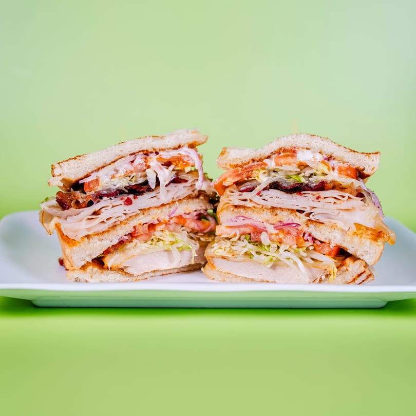 Two halves of a large, layered club sandwich with turkey, bacon, lettuce, tomato, and onion.