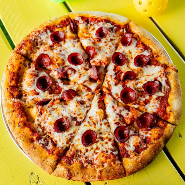 A whole pepperoni pizza from Clutch City Kitchen, sliced and ready to eat on a yellow table.