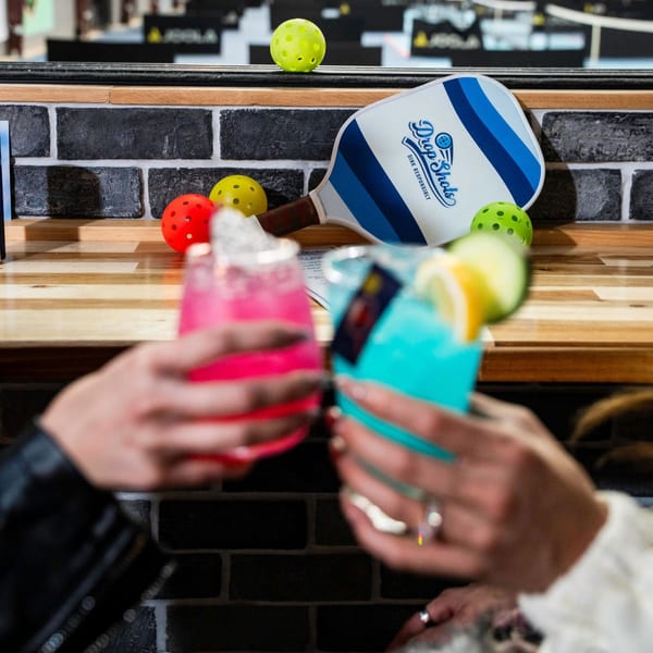 Two people clinking colorful cocktails, with pickleball paddles and balls on a brick and wood bar.