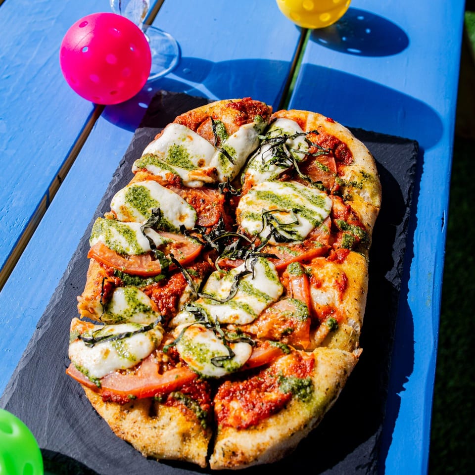 Clutch City Kitchen's flatbread pizza with pesto, mozzarella, tomatoes, and basil on a blue table.