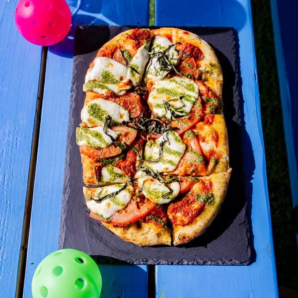 Clutch City Kitchen's flatbread pizza with mozzarella, tomatoes, pesto, and basil on a blue table.