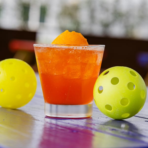 Orange cocktail with an orange slice garnish, flanked by two yellow pickleballs on a colorful table.