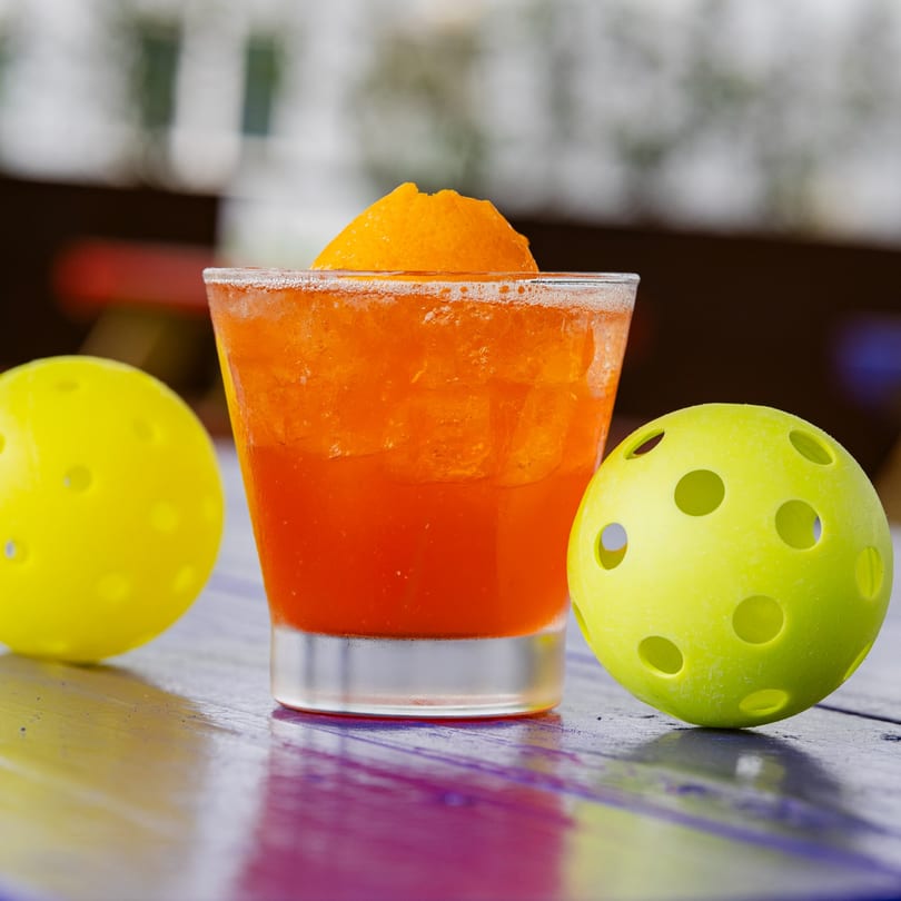 Orange cocktail with an orange slice garnish, flanked by two yellow pickleballs on a colorful table.