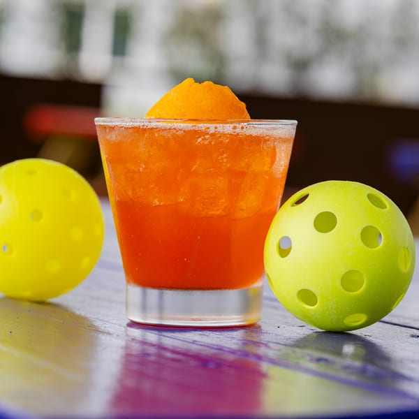 Orange cocktail with an orange slice and two yellow pickleballs on a colorful wooden table.